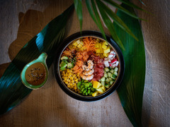 Frische Poké Bowl mit Garnelen, Mango, Edamame, Avocado, Rotkohl und Karotten, serviert mit einem Dip auf tropischen Blättern angerichtet.