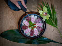 Kunstvoll angerichtetes Dessert mit rosa Blüten-Optik, serviert in einer schwarzen Schale auf tropischem Blatt – stilvoll ergänzt durch eine Orchidee und Hand mit Löffel.