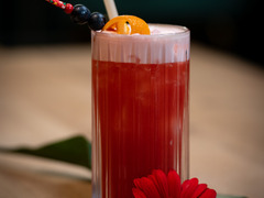Fruchtiger Cocktail mit Orangenscheibe und Cocktailkirsche, serviert im hohen Glas mit Strohhalm – dekoriert mit einer roten Blüte.