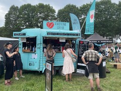 FirstLoveCoffee GmbH - Kaffee Catering Berlin - Mobiler Kaffeeanhänger auf grünem Festivalgelände mit Bio-Kaffee, Iced Latte, Iced Chai, Fahnen und Barista-Service für Sommer-Events.