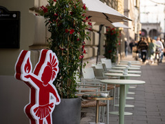 Einladender Außenbereich eines Restaurants mit Tischen, Sonnenschirmen und auffälliger roter Werbefigur – stilvolle Straßenszene mit mediterranem Flair.