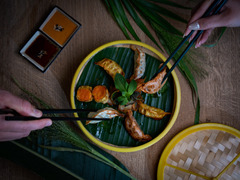 Asiatische Spezialitätenplatte mit Gyoza, Shrimps und Gemüse, serviert auf Bambusteller mit Dip und dekorativem Palmblatt – zwei Personen greifen mit Stäbchen zu.