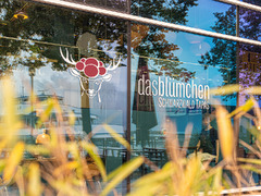 Außenansicht des Restaurants Das Blümchen Friedrichshafen mit Logo und Schriftzug „Schwarzwald Tapas“ auf der Glasfassade – moderne Architektur mit Seespiegelung.