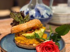 Die Feinschmecker Catering & Partyservice Hamburg - Vegetarischer Sesam-Bagel mit Hummus-Aufstrich und Avocado, stilvoll präsentiert auf einem blauen Teller neben einer roten Rose für exklusive Frühstücke und Brunches.