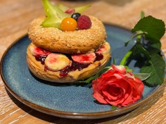 Die Feinschmecker Catering & Partyservice Hamburg - Süßer Frühstücks-Bagel mit Erdnussbutter oder Nuss-Nougat-Creme, Bananenscheiben und Beerenmarmelade, garniert mit Sternfrucht und Himbeeren neben einer roten Rose.