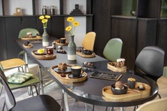 Der modern gestaltete Café-Innenbereich von Kevin Kugel Chocolatier in Sindelfingen. Zu sehen ist ein langer, dunkler Holztisch mit farbigen Designerstühlen (Grün, Gelb, Schwarz) und gelben Blumen in Vasen. Auf dem Tisch sind hölzerne Serviertabletts mit 