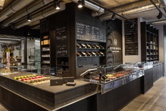 Die edle, dunkel gestaltete Verkaufstheke im Ladengeschäft von Kevin Kugel Chocolatier. Im Vordergrund stehen verglaste Vitrinen, die prall gefüllt sind mit bunten Patisserie-Törtchen und einer riesigen Auswahl handgemachter Pralinen. Im Hintergrund hänge