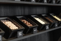 Eine hochauflösende Nahaufnahme (Makro-Produktfoto) aus dem Verkaufsregal. Gezeigt werden schwarze Präsentationsboxen, in denen feine, handgemachte Pralinen und Trüffel perfekt aufgereiht liegen. Vor den Boxen sind edle, kleine schwarze Beschriftungsschil