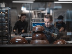 Ein exklusiver Blick hinter die Kulissen in die gläserne Schokoladenmanufaktur. Der deutsche Chocolatier-Meister Kevin Kugel arbeitet in schwarzer Meisterkleidung und Handschuhen konzentriert in der Produktion. Vor ihm auf der Arbeitsfläche befinden sich 